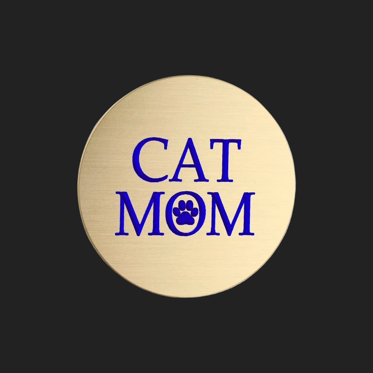 A round, brushed gold metal sticker featuring the text 'CAT MOM' in bold blue lettering, with a blue paw print icon replacing the letter 'O', set against a plain white background.