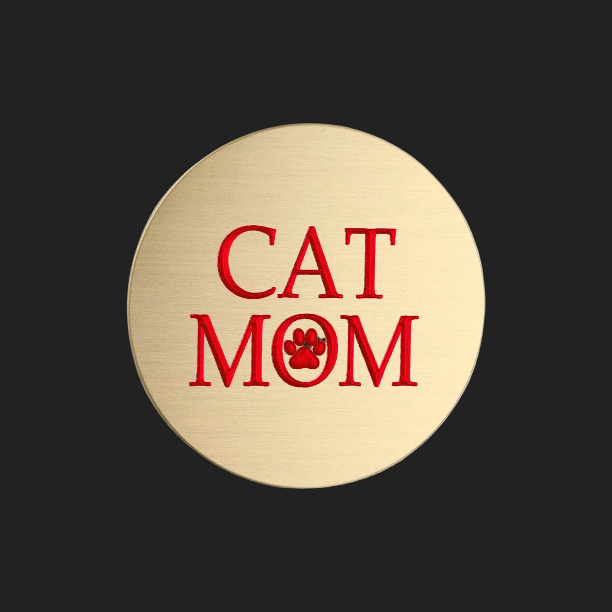 A round, brushed gold metal sticker featuring the text 'CAT MOM' in red, with the letter 'O' replaced by a red paw print icon, shown on a plain white background.