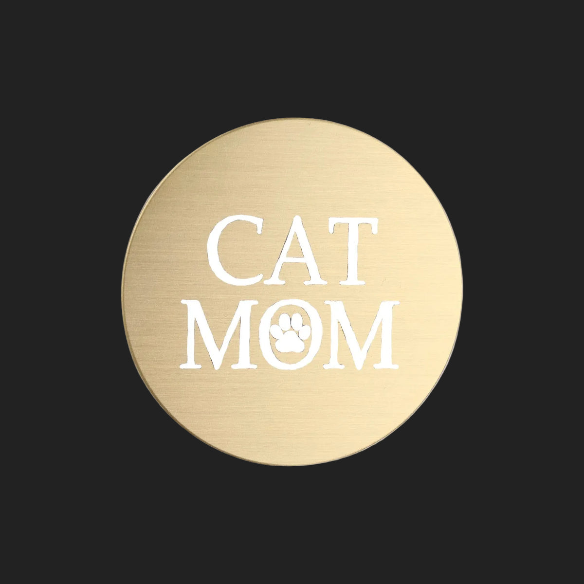 A round, brushed gold metal sticker featuring the words 'CAT MOM' in white, with a paw print icon inside the letter 'O', set against a plain white background.