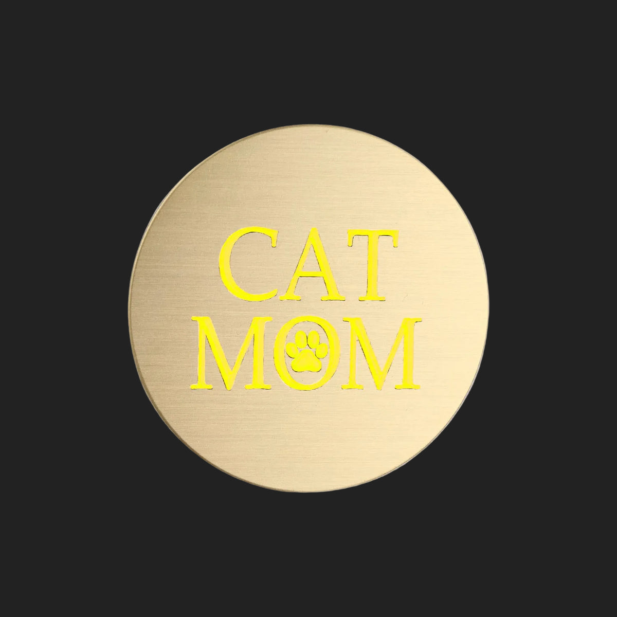 A round brushed gold metal sticker featuring the words 'CAT MOM' in bright yellow lettering, with a small yellow paw print inside the letter O, shown on a plain white background.