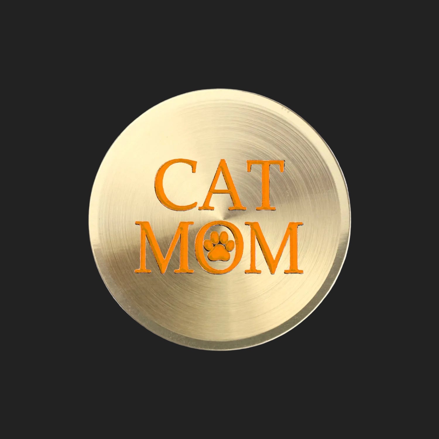 A round, brushed gold metal sign or plaque featuring the text 'CAT MOM' in orange, with the letter 'O' stylized as a paw print, set against a plain white background.