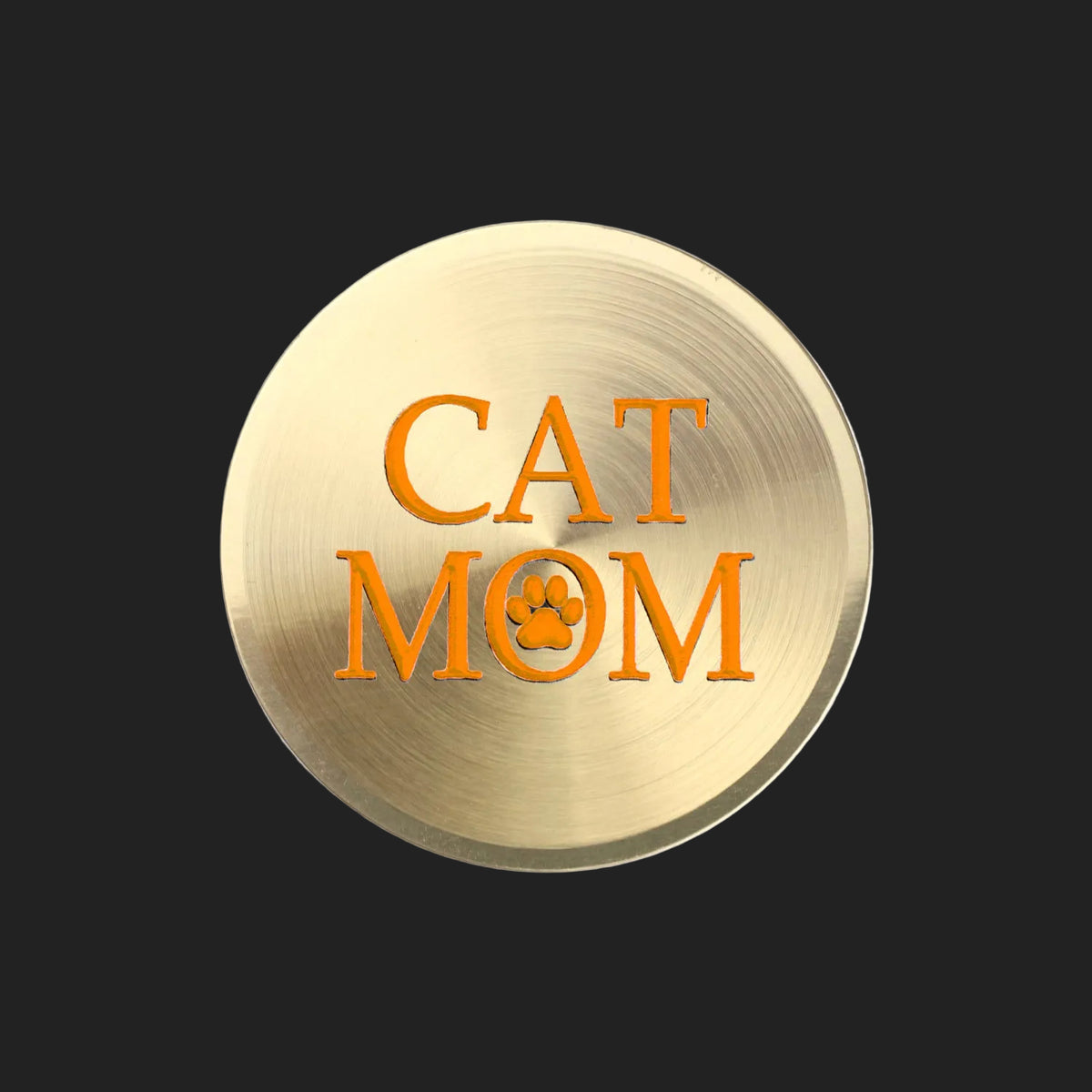 A round, brushed gold metal sign or plaque featuring the text 'CAT MOM' in orange, with the letter 'O' stylized as a paw print, set against a plain white background.
