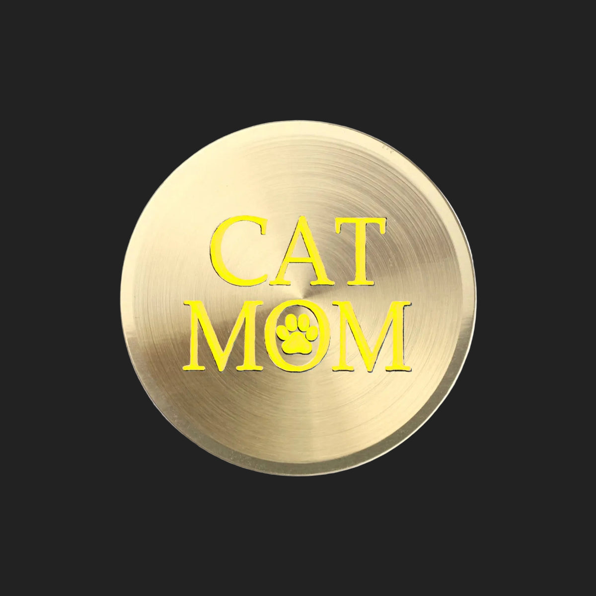 A round, brushed gold metal sticker featuring the text 'CAT MOM' in yellow, with the letter 'O' replaced by a small yellow paw print icon, shown on a plain white background.