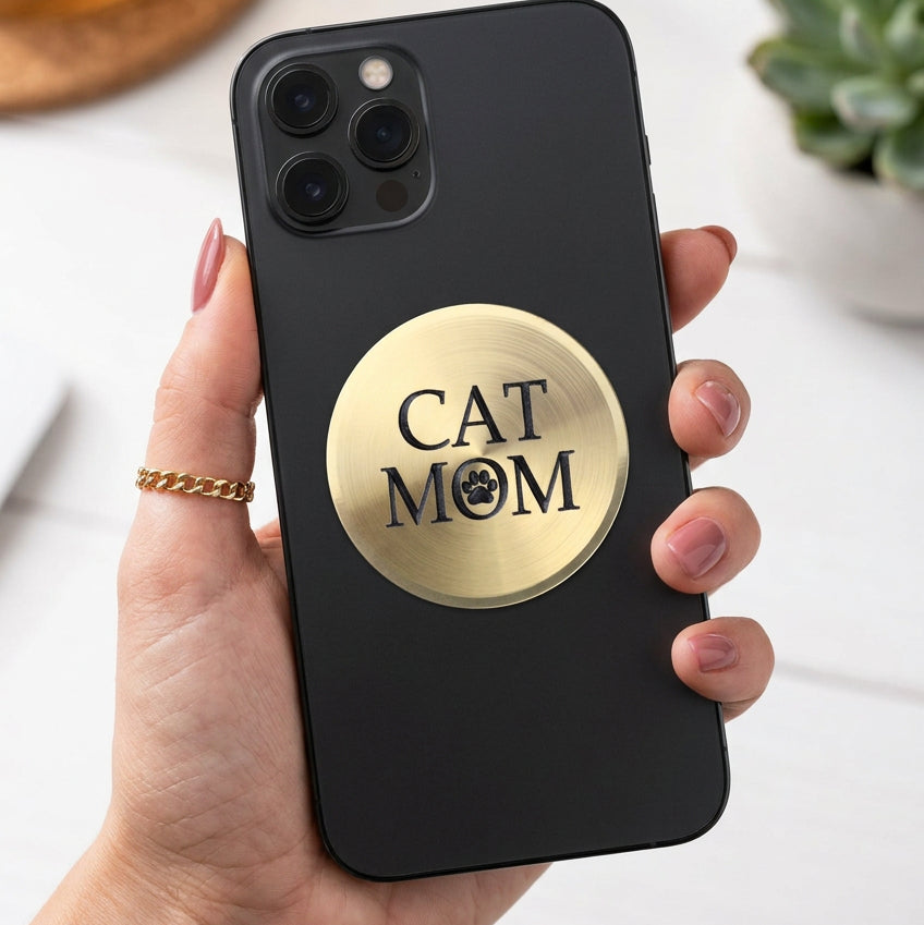 A round, brushed gold metal plaque featuring the text 'CAT MOM' in black, with the letter 'O' replaced by a black paw print icon, shown on a plain white background.