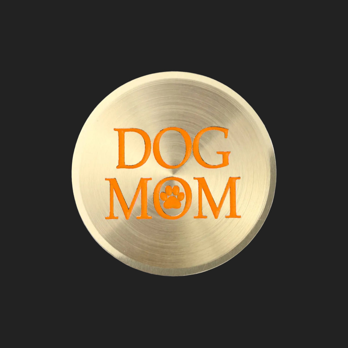 A round, brushed gold metal sticker featuring the words 'DOG MOM' in orange lettering with a paw print icon, set against a plain white background.
