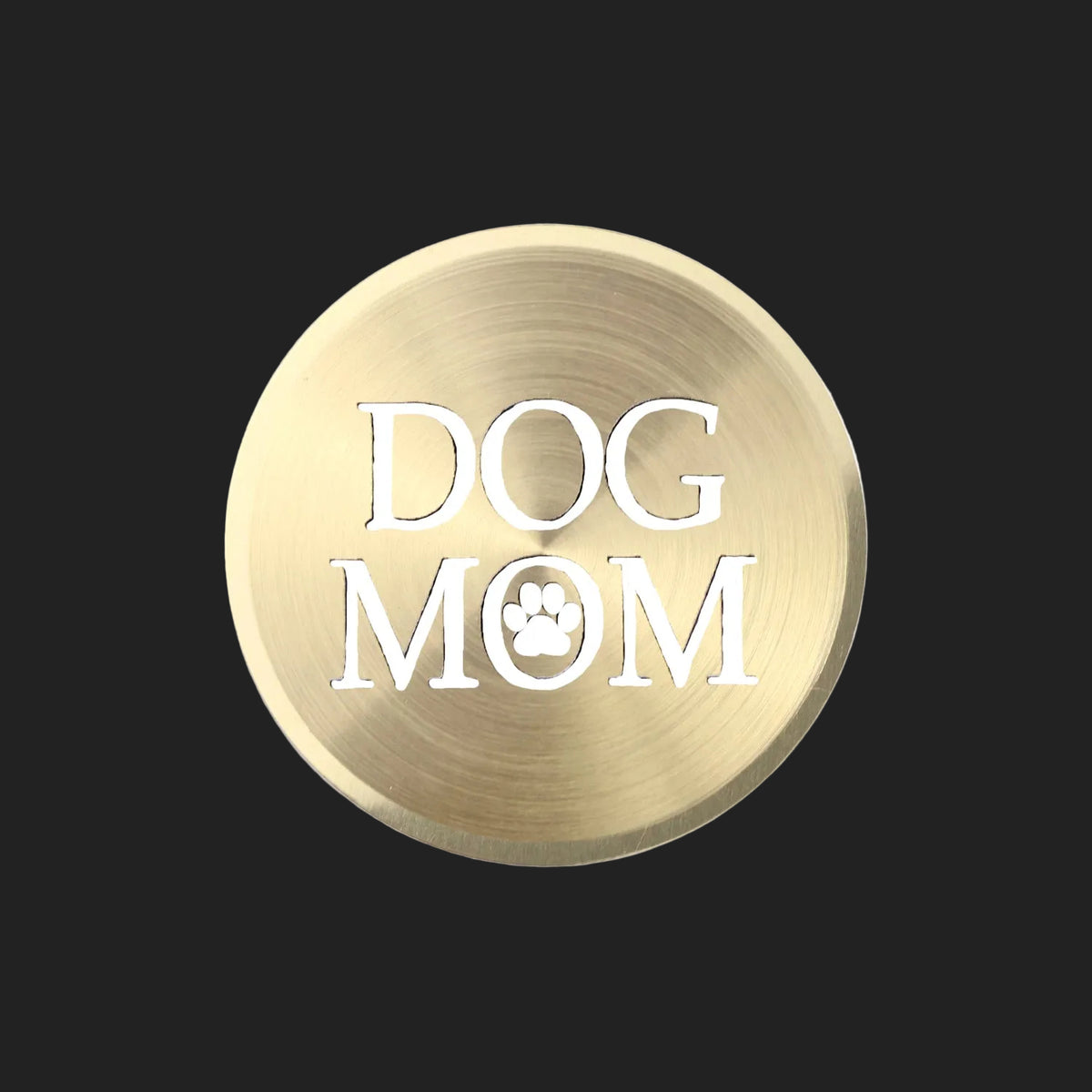 A round, brushed metallic gold sign or plaque featuring the text 'DOG MOM' in white, with a paw print icon inside the letter 'O'.