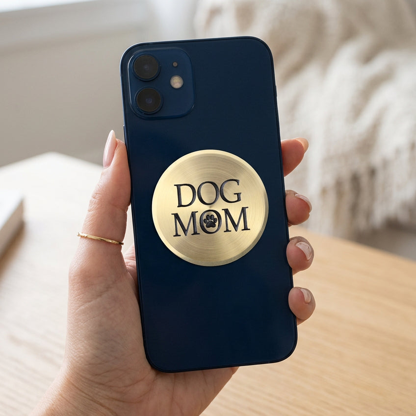 A round, brushed brass or gold-colored metal sign with the words 'DOG MOM' in black lettering and a black paw print icon, shown on a plain white background.