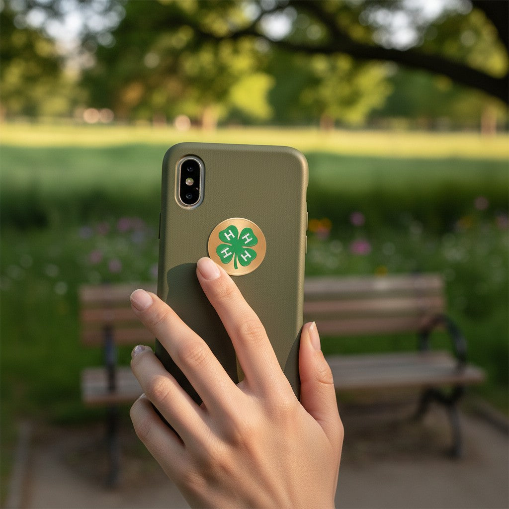 Metal Stickems four leaf clover metal sticker on an olive green phone case held in a hand in a park setting.