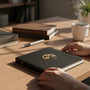 Metal Stickems 2-inch antique gold educational achievement award sticker applied to a black notebook on a desk.