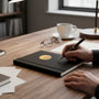 Metal Stickems gold apple and books emblem adhered to a black notebook on a wooden desk with a hand writing with a pen