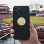 Metal Stickems gold baseball emblem sticker applied to a black smartphone case at a baseball stadium.