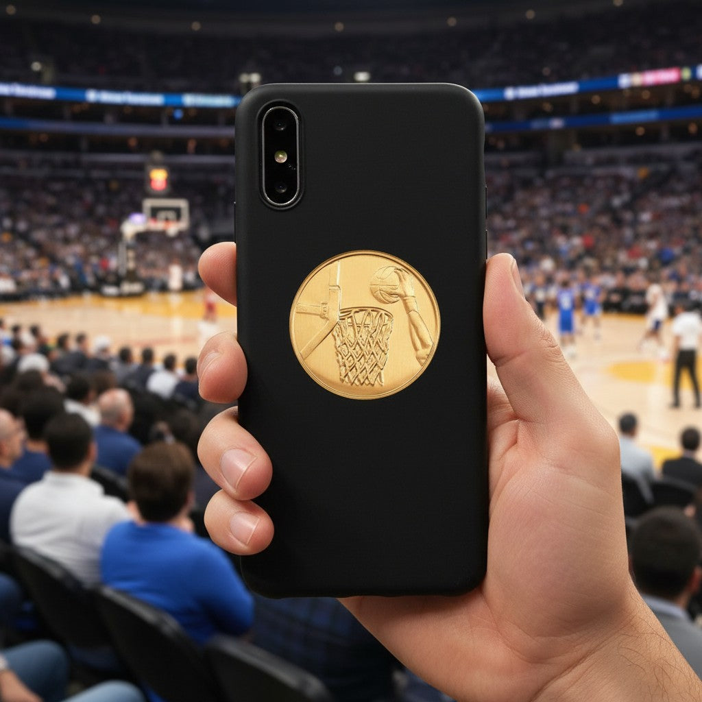 Metal Stickems gold basketball hoop emblem sticker applied to a black smartphone case in a stadium setting.