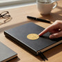 Metal Stickems gold metal lamp and book emblem sticker applied to a black notebook on a wooden desk.