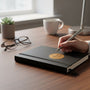 Metal Stickems 2-inch gold commemorative real estate closing coin sticker applied to a black notebook on a desk.