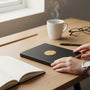 Metal Stickems gold shooting sports emblem sticker featuring crossed rifles on a black notebook on a wooden desk.