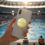 Metal Stickems 2-inch gold swimming achievement emblem applied to the back of a smartphone at a swimming pool.