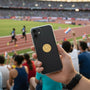 Metal Stickems gold embossed male track events sticker on a black smartphone held at a stadium track meet.