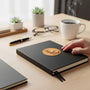 Metal Stickems gold praying hands metal sticker applied to a black notebook on a desk with coffee and glasses.