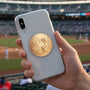 Metal Stickems round gold baseball sticker with embossed player design on a smartphone case at a stadium.